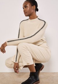Beige gerippter Strickpullover mit schwarzem Stitching an den Ärmeln, kombiniert mit beigen Hosen mit Bündchen-Details und schwarzen Stiefeletten.