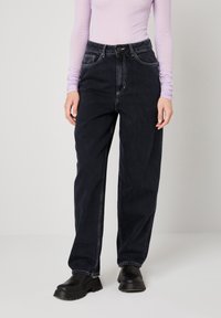 Osoba odjevena u crne traperice visokog struka širokih nogavica, crne masivne cipele i ljubičastu majicu dugih rukava, stoji na bijelom podu.