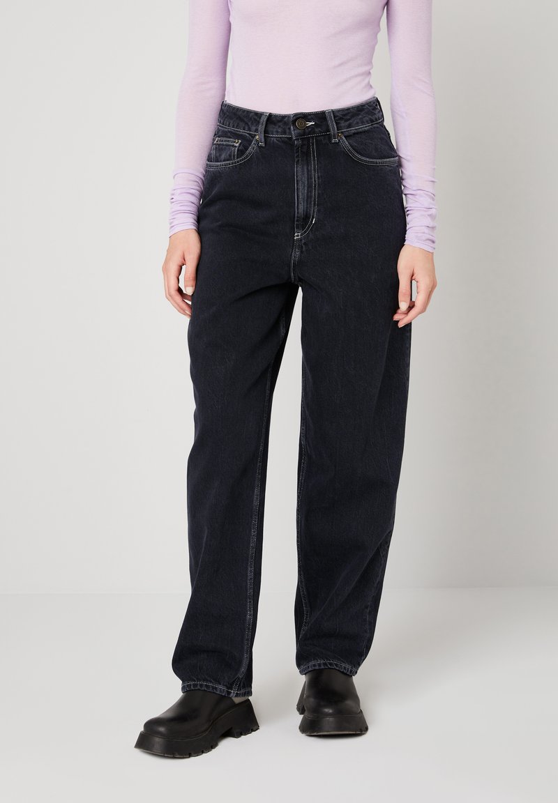 Osoba odjevena u crne traperice visokog struka širokih nogavica, crne masivne cipele i ljubičastu majicu dugih rukava, stoji na bijelom podu.