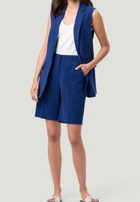 Blazer senza maniche di colore blu navy abbinato a un top bianco e pantaloni corti blu navy, con tasche e una vestibilità sartoriale, realizzato in tessuto leggero.