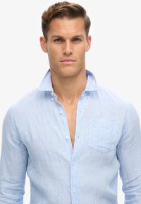 Camisa de botones de manga larga en azul claro con cuello clásico, que presenta un bolsillo en el pecho y sutiles rayas verticales a lo largo.