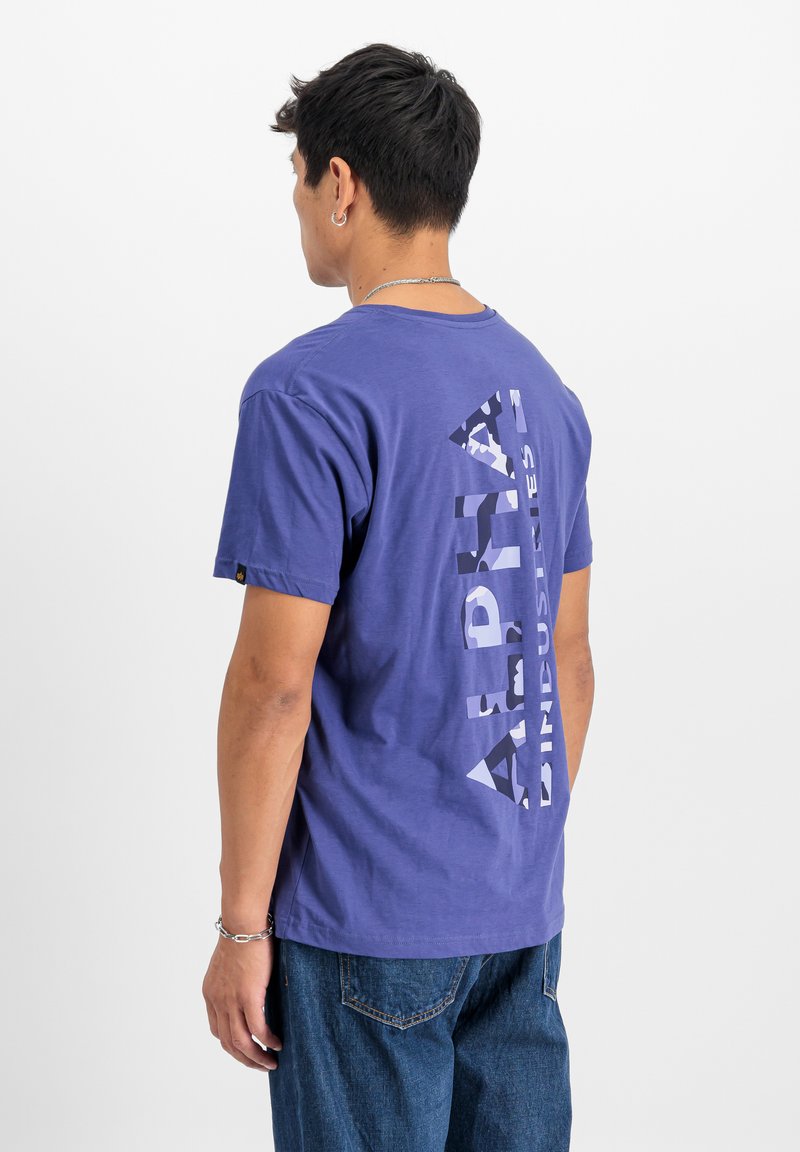 Violeta kokvilnas T-kreklu ar lielu, vertikāli izkārtotu alfas grafiku kamuflāžas drukā. Īsām piedurknēm un apaļu kakla izgriezumu.