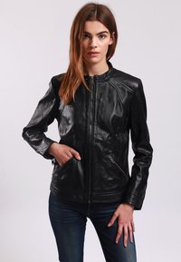 Giacca di pelle nera con colletto alto, chiusura a zip e due tasche laterali con zip. Presenta una superficie liscia e un design aderente.