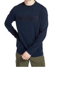 Hombre con sudadera azul marino de Timberland con logo y texto, combinada con pantalones cargo verde oliva, con las manos resting cerca de la cintura.