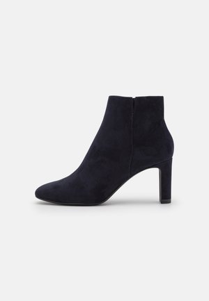 Bottine noire en daim avec un bout pointu, un talon bloc de hauteur moyenne et une fermeture éclair latérale sur fond blanc.