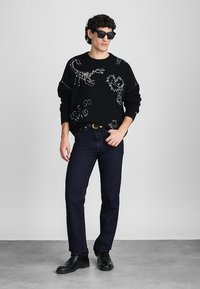 Black knitted sweater with white dinosaur designs, paired with dark blue jeans and black shoes. Model wearing sunglasses, standing against a neutral background.