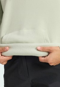 Zielona bluza z miękkim, ocieplanym dołem i przednią kieszenią, trzymana w dłoniach. Posiada gładką teksturę i prosty design.