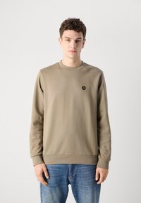 Beige long-sleeve sweatshirt with a crew neck and ribbed cuffs. Features a small black logo on the chest. Paired with blue denim jeans.
