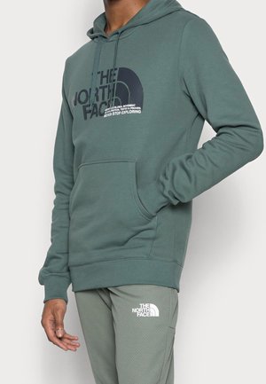 Mężczyzna ubrany w zieloną bluzę The North Face z kapturem i kieszenią z przodu oraz pasujące zielone spodnie z logo The North Face na udzie, stojący na białym tle.