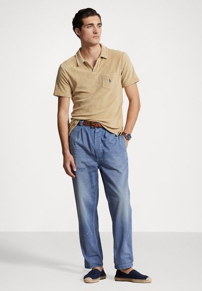 Polo shirt beige avec une poche et un logo bleu, associé à un pantalon bleu clair légèrement usé et des espadrilles navy. Ceinture tressée marron.