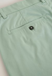 Poche arrière d'un pantalon vert clair avec fermeture à bouton et passants de ceinture, montrant des coutures détaillées et la texture du tissu.