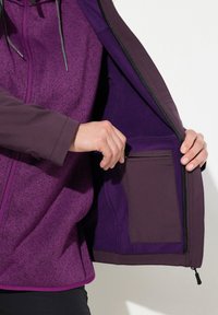 Chaqueta morada con cremallera, exterior de tela texturizada, forro interior suave y un pequeño bolsillo interior. Presenta un dobladillo curvado y capucha ajustable.