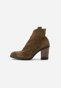 Stivale alla caviglia in suede marrone con punta arrotondata, pannello elastico laterale e un tacco medio in legno. Texture liscia, design semplice, forma versatile.