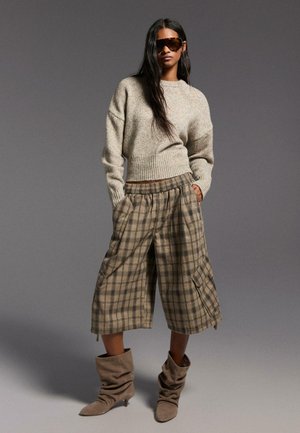 Femme portant un pull beige oversize, des culottes larges à carreaux, des bottes en daim décontractées et de grandes lunettes de soleil foncées sur un fond gris uni.
