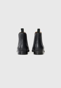 Botas de tobillo de cuero negro con una textura suave, con un tirador verde y una punta redondeada, descansando sobre un tacón bajo y robusto de color negro.