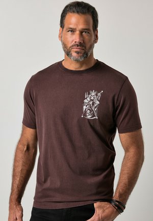 Mand med skæg iført en brun t-shirt med en hvid skeletkonge grafik på venstre bryst, stående mod en ensfarvet baggrund.