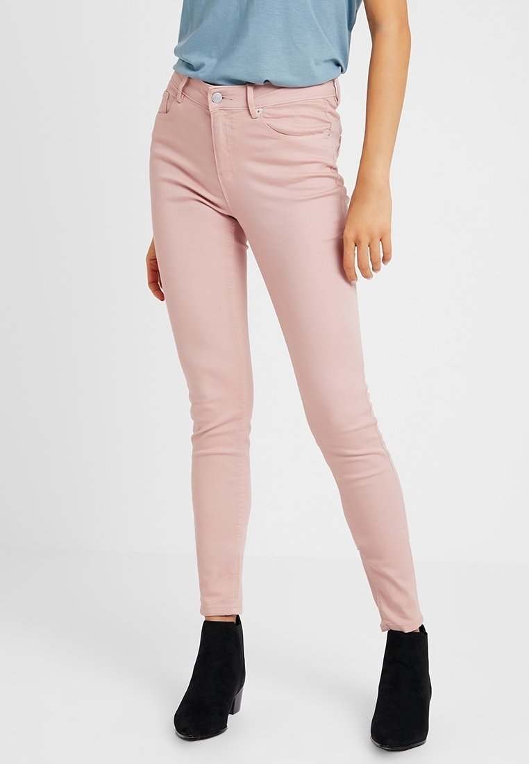 Femme portant un jean skinny rose clair, des bottines noires, et une chemise bleue rentrée, debout devant un fond blanc uni.
