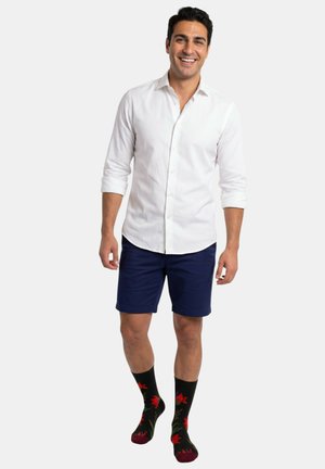 Homme souriant portant une chemise blanche à boutons avec les manches retroussées, un short bleu marine et des chaussettes noires à motifs floraux rouges.