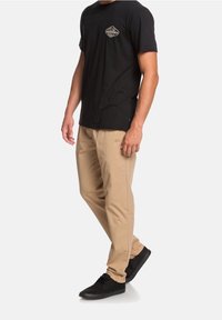 Svart bomullströja med en liten logotyp på bröstet, kombinerad med beige byxor och svarta sneakers, som visar en avslappnad stil och neutrala färger.