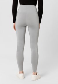 Leggings a vita alta di colore grigio chiaro, realizzati in un materiale morbido e elastico; presentano un rinforzo arrotondato e un design aderente, indossati con sneakers bianche.