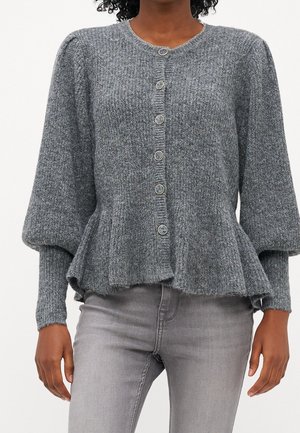 Kvinde iført en grå cardigan med knapper, puffede ærmer og en flæsekant, sammen med lysegrå jeans mod en ensfarvet baggrund.