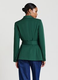 Blazer verde su misura con vita rivestita, spalle strutturate e maniche lunghe eleganti; abbinato a pantaloni grigio scuro texturizzati.
