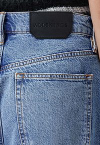Světle modré džíny z denimu s černou koženou nášivkou s vyraženým textem. Oranžové švy zdobí kapsy a švy.