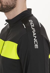 Schwarze Jacke mit einem gelben horizontalen Streifen. Verfügt über einen hohen Kragen, einen Reißverschluss und den weißem "ENDURANCE"-Text auf der Schulter.