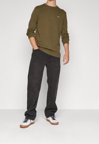 Oliivinvihreä neulepaita, jossa on resorit hihansuissa, yhdistetty tummanharmaisiin farkkuihin ja valkoisiin tennareihin, joissa on siniset nauhat ja ruskeat pohjat.