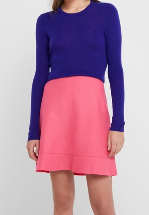 Femme portant un haut royal bleu ajusté à manches longues et une jupe évasée rose taille haute, debout devant un fond uni.