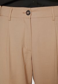 Pantalones beige con un botón negro abrochado en la cintura, con trabillas para cinturón y costuras visibles en los paneles frontales.
