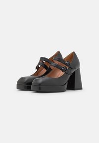 Black leather platform heels with wide block heel, circular buckle straps, rounded toe, and smooth texture. Interior is brown.
