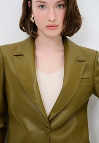 Blazer in pelle verde oliva con design strutturato, revers a punta, chiusura con un bottone e taschino sul petto, indossato sopra un top beige.