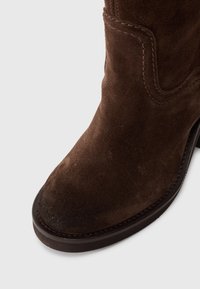 Botte cheville en daim marron avec un bout arrondi, des coutures décoratives et une semelle contrastante. Présente une texture lisse et un léger talon.