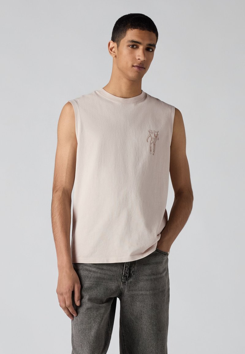 Giovane uomo che indossa una maglietta chiara senza maniche con una piccola stampa sul petto e jeans grigio scuro, posando con una mano in tasca su sfondo semplice.
