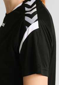 Svart idrottströja med mesh-detaljer på axlarna, kontrasterad av vita och mönstrade inslag. Slät textur, figursydd passform.