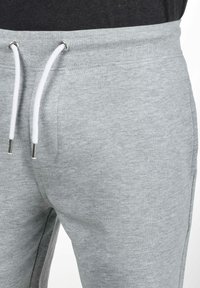 Pantalon de survêtement gris en matériau doux avec une taille côtelée, doté d'un cordon de serrage blanc avec des embouts en métal argenté.