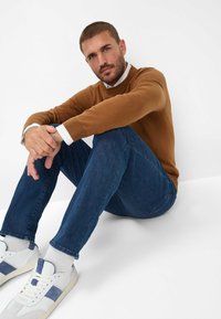 Maglione marrone, camicia bianca sotto, jeans blu, sneakers bianche con accenti blu. Uomo seduto, posizione rilassata su uno sfondo bianco.