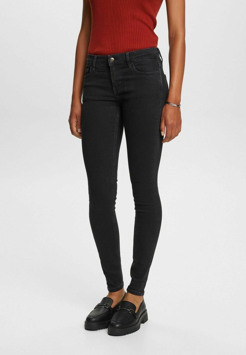 Femme portant un jean skinny noir, des mocassins noirs et un top rouge sans manches côtelé, debout sur un fond blanc.