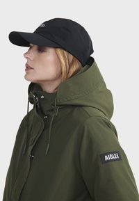 Veste imperméable verte avec un col haut, cordons de serrage réglables et un patch logo noir. Associée à une casquette noire avec une visière incurvée.