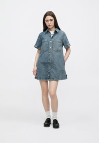Φόρεμα από τζιν με κοντά μανίκια σε ανοιχτό μπλε, με δύο τσέπες στο στήθος, κουμπιά μπροστά και ξεφτισμένο τελείωμα· φοριέται με μαύρα παπούτσια και γκρι κάλτσες.
