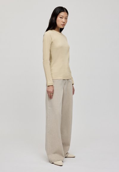 Langärmliges, beiges Shirt kombiniert mit lockeren, hellbeigen, hochtaillierten Hosen. Glatte Textur mit entspannter Passform. Spitz zulaufende cremefarbene Schuhe.