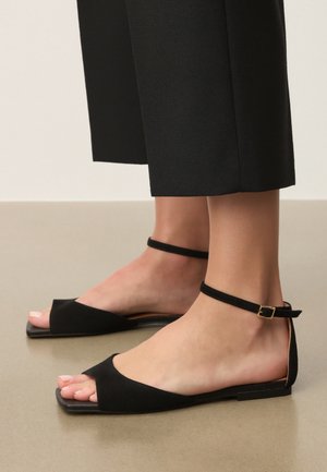 Fødder iført sorte flade sandaler med ankelremme og åbne tæer, kombineret med korte sorte bukser på en beige gulv.