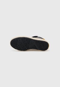 Semelle de sneaker noire avec motif en caoutchouc texturé, dotée d'une intercalaire beige et d'un accent jaune. Matière supérieure lisse avec une sangle à bande auto-agrippante.