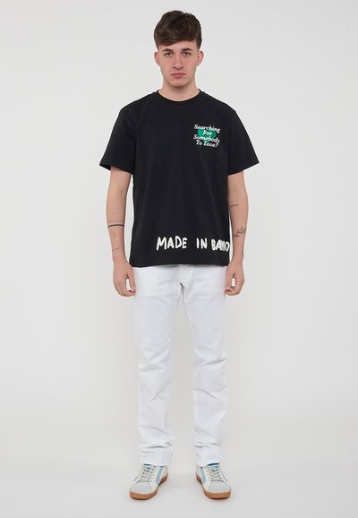 T-shirt di cotone nera con testo verde che recita "Cercando Qualcuno Da Amare?" e stampa bianca "FATTO A BALI"; abbinata a jeans bianchi e sneakers.