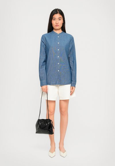 Donna che indossa una camicia azzurra con bottoni, pantaloni corti bianchi, scarpe bianche a punta, mentre tiene una borsa nera a tracolla su uno sfondo neutro.