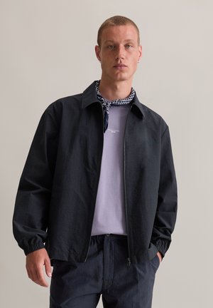 Zwarte lichtgewicht jack met een ritssluiting aan de voorkant, elastische manchetten en een kraag, gedragen over een lavendelkleurig shirt met een klein logo en een gepatterde sjaal.