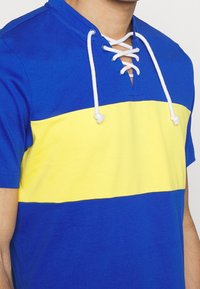 Modrobíložluté tričko s krátkými rukávy a šněrovacím výstřihem. Má vodorovné pruhy a měkkou, hladkou texturu látky.