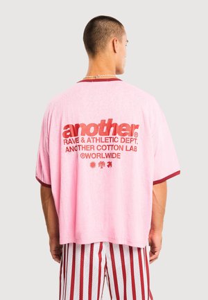 Man wearing a loose pink T-shirt with red text and symbols on the back, paired with red and white vertical striped shorts.