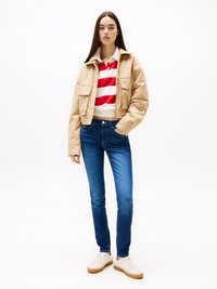Tan geknipte jas, rood-witte gestreepte polo, donkerblauwe skinny jeans en witte sneakers met een beige zool. Model staat voor een effen achtergrond.